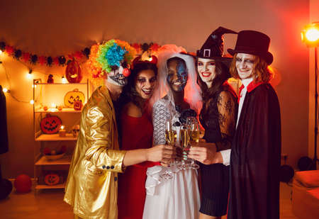 Diverse group of happy young people drinking and having fun at Halloween party at home. Smiling adult multiethnic male and female friends in scary costumes standing together and clinking flute glassesの写真素材