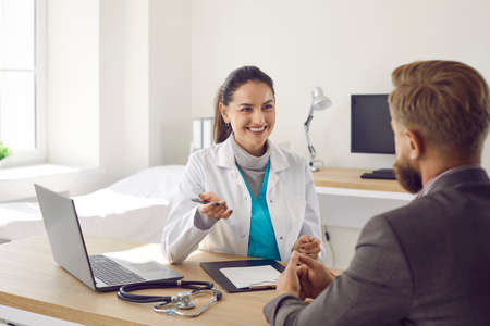 Happy friendly female doctor tells her patient good news during meeting in her office. Unidentified male patient sitting with his back to camera talking to his family doctor. Healthcare concept.の写真素材