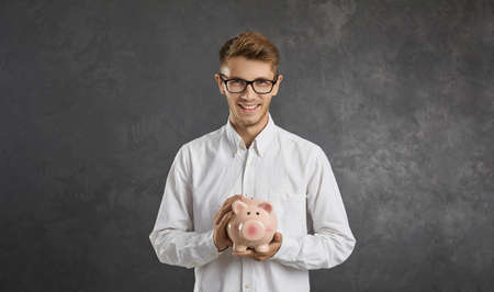 Save money. Studio portrait of smiling successful young man who is holding piggy bank standing on gray background. Concept of investment, loans, deposits and finance, wealth and accumulation.の写真素材