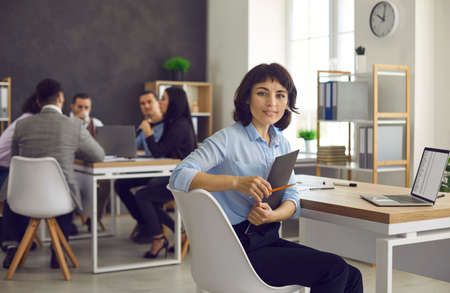Beautiful young European woman sitting at desk in office workspace, holding presentation notes and looking at camera. Portrait of professional business executive or company worker in her workplaceの写真素材