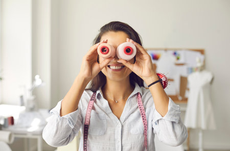 Portrait of cheerful funny seamstress looking through spools of thread as if through binoculars. Woman with pin pad on her arm and with measuring tape around her neck is having fun in sewing studio.の写真素材