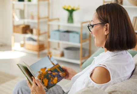 Happy woman in glasses sitting on sofa at home and looking through pictures in printed family album....