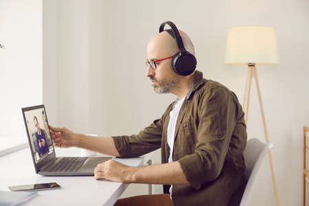 Serious focused bald man wearing headphones and glasses sitting at modern laptop in home office,...