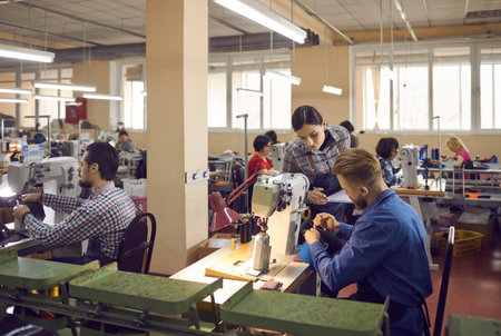People working in a big workshop room at a shoe factory. Male and female workers sitting at tables with industrial sewing machines and making new footwear details. Manufacturing industry conceptの写真素材