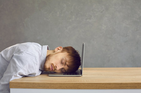 Tired male office worker sleeping sitting at a desk putting his head on a laptop keyboard. Side view of a young man on a gray background with space for text. Concept of burnout, fatigue and laziness.の写真素材