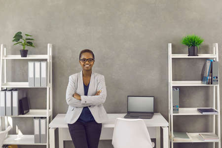 Portrait of happy business lady at work. Beautiful young black woman in suit and glasses leaning on...