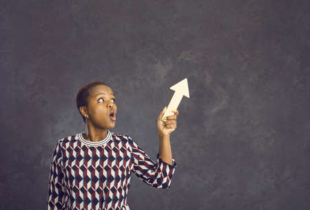 Shocked young african american woman with wooden arrow pointing up standing on gray background....