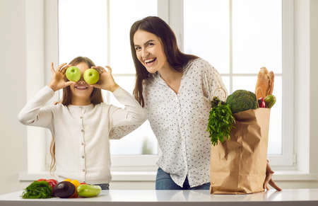 Happy girl covering eyes with green apples to make crazy funny face. Portrait of cheerful family having fun while unpacking groceries at kitchen table with paper bag full of natural vegetarian foodsの写真素材