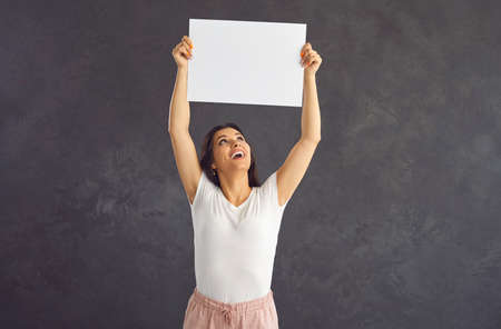 Young beautiful woman over her head holding blank white sheet of paper standing on gray background. Caucasian brunette woman in casual clothes looks up contentedly. Mockup, layout, copy space.の写真素材