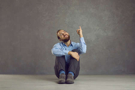 Funny caucasian man thinking sitting with raised finger on the floor on gray background. Smiling...