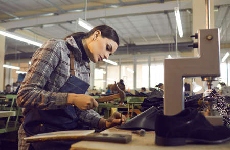 Female worker using professional tools while making new leather boots. Beautiful young woman working with hammer and nail at shoe factory workshop. Manufacturing industry, footwear production conceptの写真素材