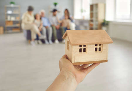 Close up real estate agents hand holding miniature toy house model against blurred background of living-room with young parents, children and grandparents all together. Family buying new home conceptの写真素材