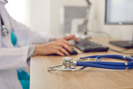 Crop close up of woman doctor sit at desk work on computer in clinic or hospital. Focus on stethoscope phonendocsope on GP table in cabinet. Medical service, healthcare and medicine concept.の写真素材