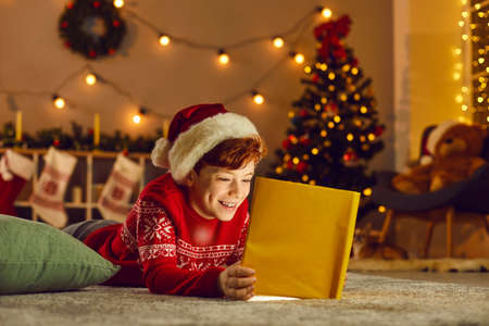 Christmas miracle. Happy boy lies on the floor in a cozy room and reads fairy tales looking at a book that glows with a magical light inside. Concept of a happy childhood. Christmas background.の写真素材