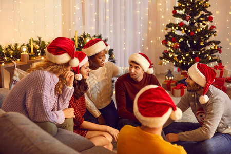 Celebration Christmas and New year. Cheerful multiracial friends communicate with each other while enjoying Christmas Eve. Young people in santa hats sit in friendly circle in living room at home.の写真素材