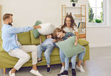 Young family of four having fun on sofa at home. Happy excited mom, dad and two children having a pillow fight and laughing sitting on couch in modern cozy Scandinavian living room interiorの写真素材