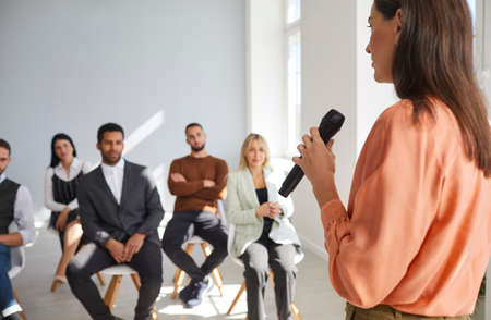 Female speaker with microphone in her hand speaks in front of small group of business people. Group of diverse people listen to their colleague at business meeting. Concept of business training.の写真素材