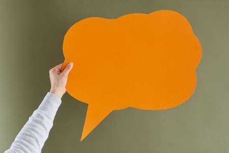 Anonymous adult female showing one orange cloud shaped mock up copyspace speech balloon or message bubble on greyish green background. Hand holding paper with free, clean space for expressing opinionの写真素材