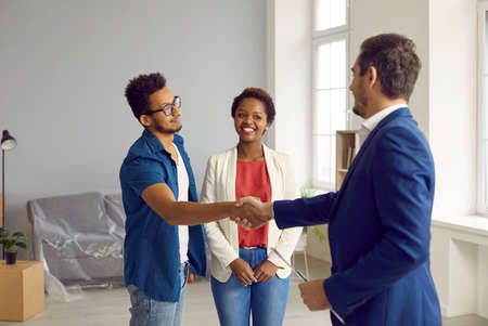 New house, new beginnings. Happy young married couple buys house and closes deal with realtor with handshake. African American family gratefully shakes hands with Caucasian real estate agent.の写真素材