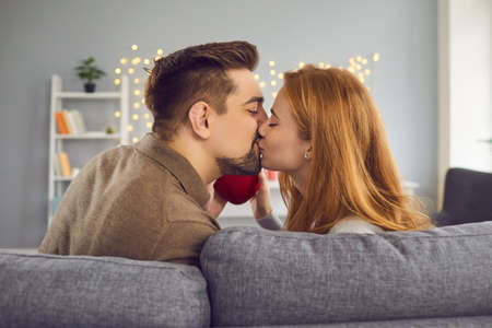 Beautiful young couple at home. Rear view of a young couple in love kissing and enjoying spending time celebrating Valentines Day with a red heart in their hand. Concept of a happy relationship.の写真素材