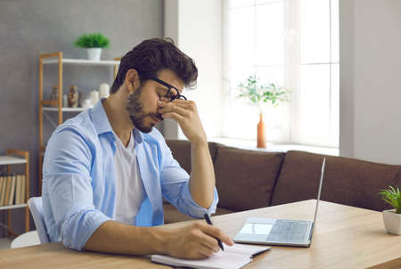 Overworked young guy sitting at desk in home office and rubbing his tired dry eyes. Man who has eye strain from constant work on laptop computer takes off eyeglasses and closes irritated eyesの写真素材