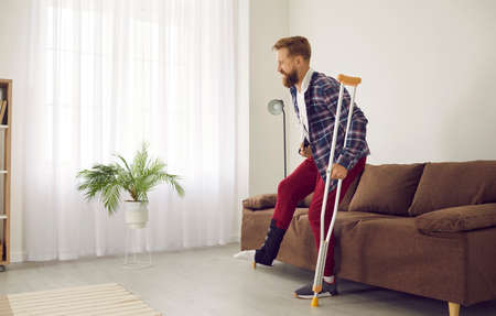 Man with leg injury leans on crutch and has difficulty getting up from couch at home. Side view of adult man with broken limb wearing bandage on his leg and having difficulty walking.の写真素材