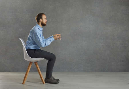 Side view portrait of a serious businessman sitting tense at an imaginary desk with a computer. Man keeps his hands outstretched as if typing on a keyboard and working on a laptop. Gray background.の写真素材