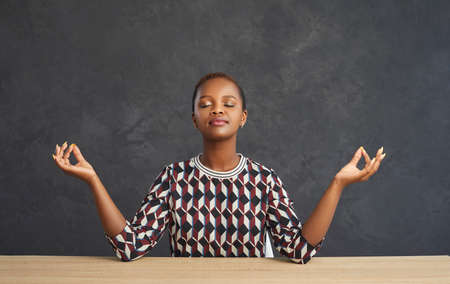 Peaceful young african american woman practicing breathing yoga exercises while sitting at the table. Happy woman sitting with closed eyes and doing relaxing meditation. Gray background.の写真素材