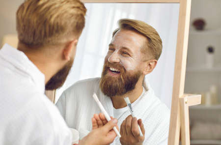 Close up mirror reflection of satisfied adult handsome bearded man who enjoys facial treatment. Cheerful man in white bathrobe uses brush to apply cosmetic mask, cream or lotion on his face.の写真素材