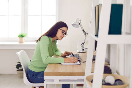 Millennial girl student sit at desk study online on computer at home. Young Caucasian woman work distant on PC, make note write in notebook. Web education, technology, freelance concept.の写真素材