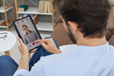 Man on digital tablet looks at profile photo of attractive young woman in dating app and presses red heart like button. Concept of finding love online. Close-up, selective focus.の写真素材