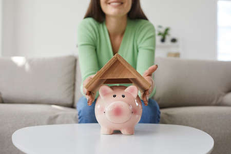 Woman saving up money to buy her own house. Happy young girl holding roof above small pink piggy bank placed on table in living room, close up. Finance, property purchase, taking mortgage loan conceptの写真素材