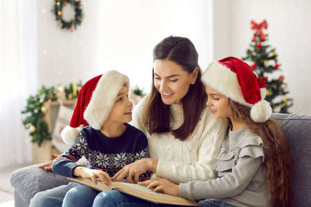 Happy family reading book on Christmas Eve at home. Mother and two children in cute red Santa hats sitting on sofa in cozy living room with Xmas tree and enjoying interesting fairytale about miraclesの写真素材