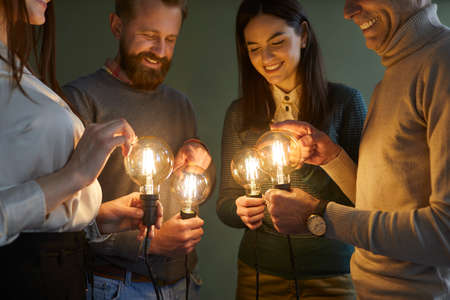 Team of people developing and trying out innovative ideas. Group of four clever men and women holding bright, shining, glowing Edison light bulbs to illuminate concept of creative idea and innovationの写真素材