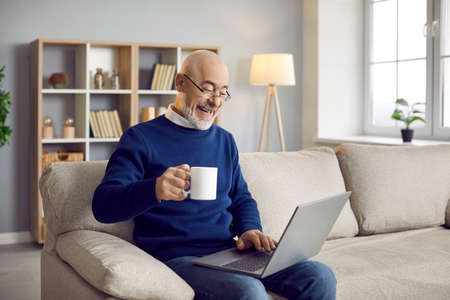 Happy cheerful bald bearded senior man enjoying leisure time on sofa in living room, holding coffee, watching comedy film on modern laptop computer device, talking to family on videocall and laughingの写真素材
