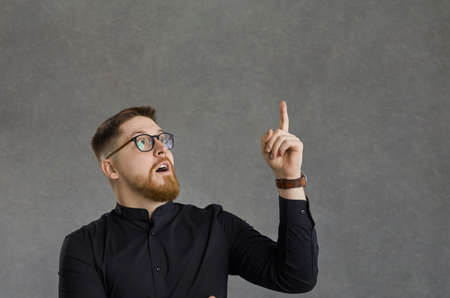 Smart excited caucasian man pointing finger up having great idea studio shot on grey background. Young guy thinking showing eureka gesture with hand upwards. Bright mind and understandingの写真素材