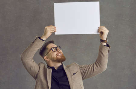 Happy smiling young man in suit and glasses showing empty white paper banner standing isolated on grey background. Studio portrait cheerful bearded hipster businessman holding clean blank mockup signの写真素材