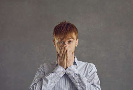 Oops. Close up portrait of attractive man with closed mouth, hands on face and eyes wide open, looking at the camera, he cannot believe his eyes, standing against gray backgroundの写真素材