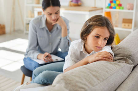 Frustrated little child at psychologists office. Portrait of girl with sad face sitting on couch during therapy session. Therapist trying to help unhappy, resentful kid who has behaviour issuesの写真素材