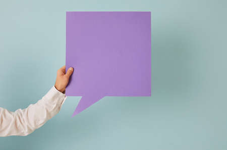Anonymous adult male holding in hand one square shaped purple mockup copyspace speech bubble on blue background. Man showing violet message balloon with free, blank, clean space for expressing opinionの写真素材
