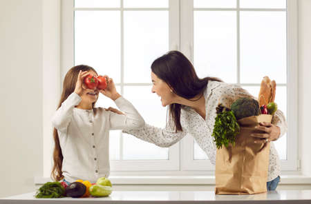 Happy little daughter covers eyes with red tomatoes and makes crazy funny face while unpacking groceries with her mom at kitchen table with paper bag full of natural vegetarian foods for family dinnerの写真素材