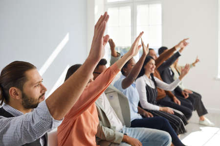 Diverse mixed race male female audience raising hands. Active multiracial multiethnic men and women sitting in row with hands up to ask coach questions after interesting talk or engaging master classの写真素材