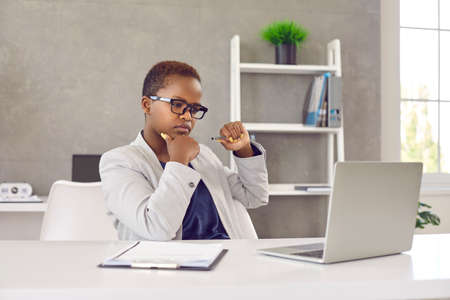 Short haired black woman in glasses sitting at working table in office, looking at laptop computer screen with pensive face expression, reading article, doing fact checking, thinking, considering ideaの写真素材