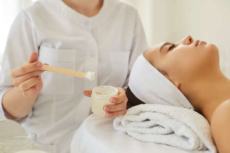 Female cosmetologist stirs and prepares moisturizing cosmetic mask to apply it to clients skin. Close up of jar with white mask or cream in hands of beautician sitting near head of female client.の写真素材