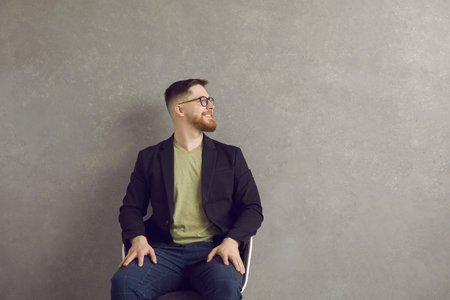 Happy cheerful man sitting on chair near grey wall, looking away at copy space background and smiling. Positive young businessman, hipster or student in glasses looking at something on the right sideの写真素材