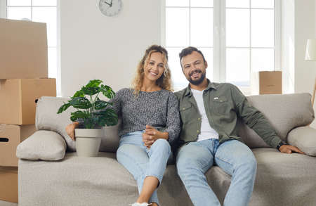 Purchase of new housing. Portrait of young happy couple sitting with green flowerpot on sofa in new...
