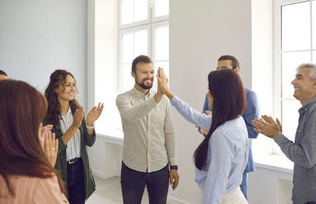Happy man and woman giving each other five to applause of colleagues after seminar training. Contact group of people standing in circle in bright room. Business training and team building concept.の写真素材