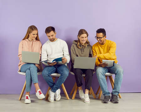 Happy multiracial college students sitting on chairs by purple wall using laptop computer devices,...