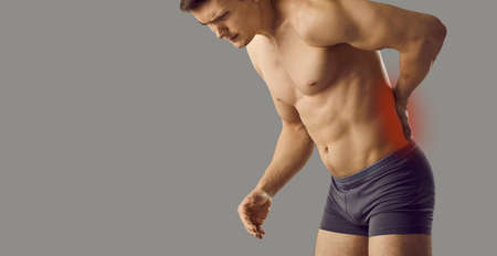Young man who has backache touching his lower back. Athletic guy in underwear isolated on grey...