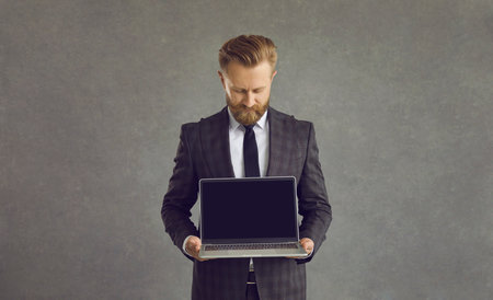 Studio shot of businessman in formal suit and tie holding showing laptop black screen blank screen monitor for text standing over grey background. Achievement, career, wealth and business conceptの写真素材